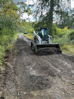 Skid steer smoothing a long gravel driveway through the woods